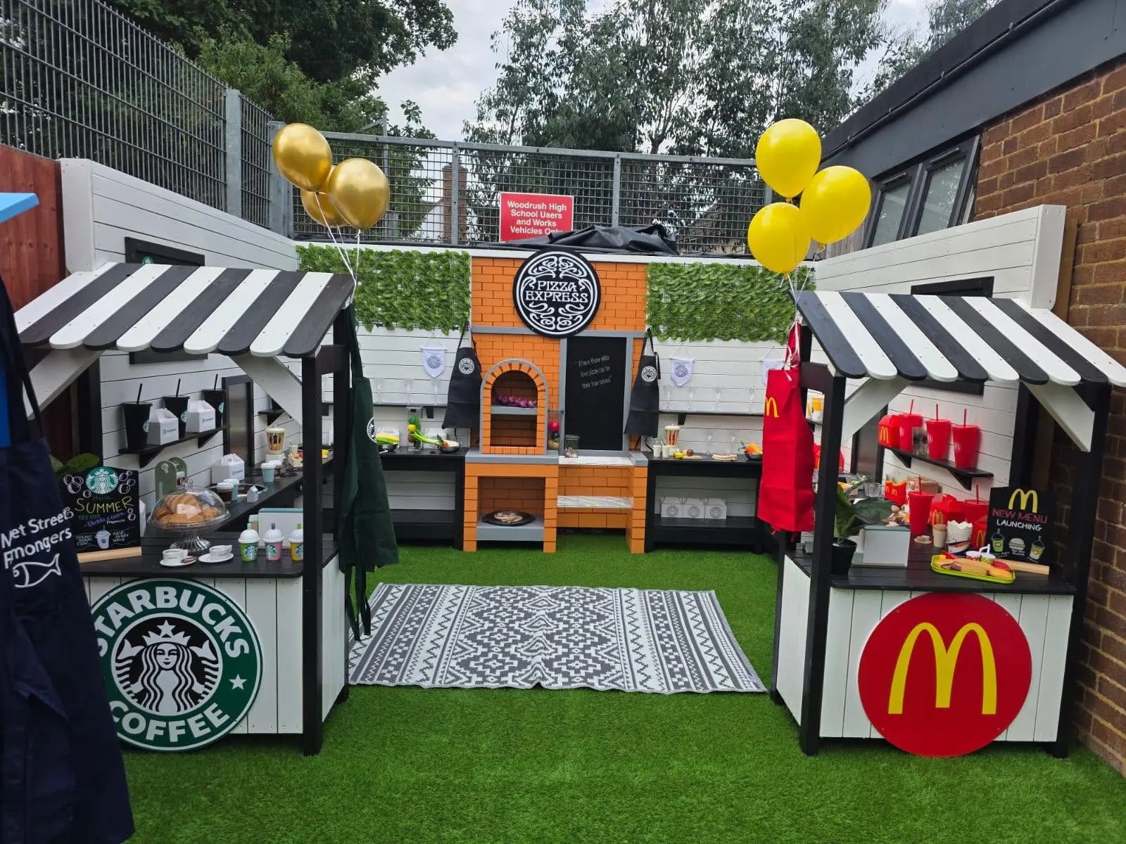 The outdoor food court at Mini Minds Play Village — a pretend coffee stall, burger bar and brick pizza kitchen on artificial grass