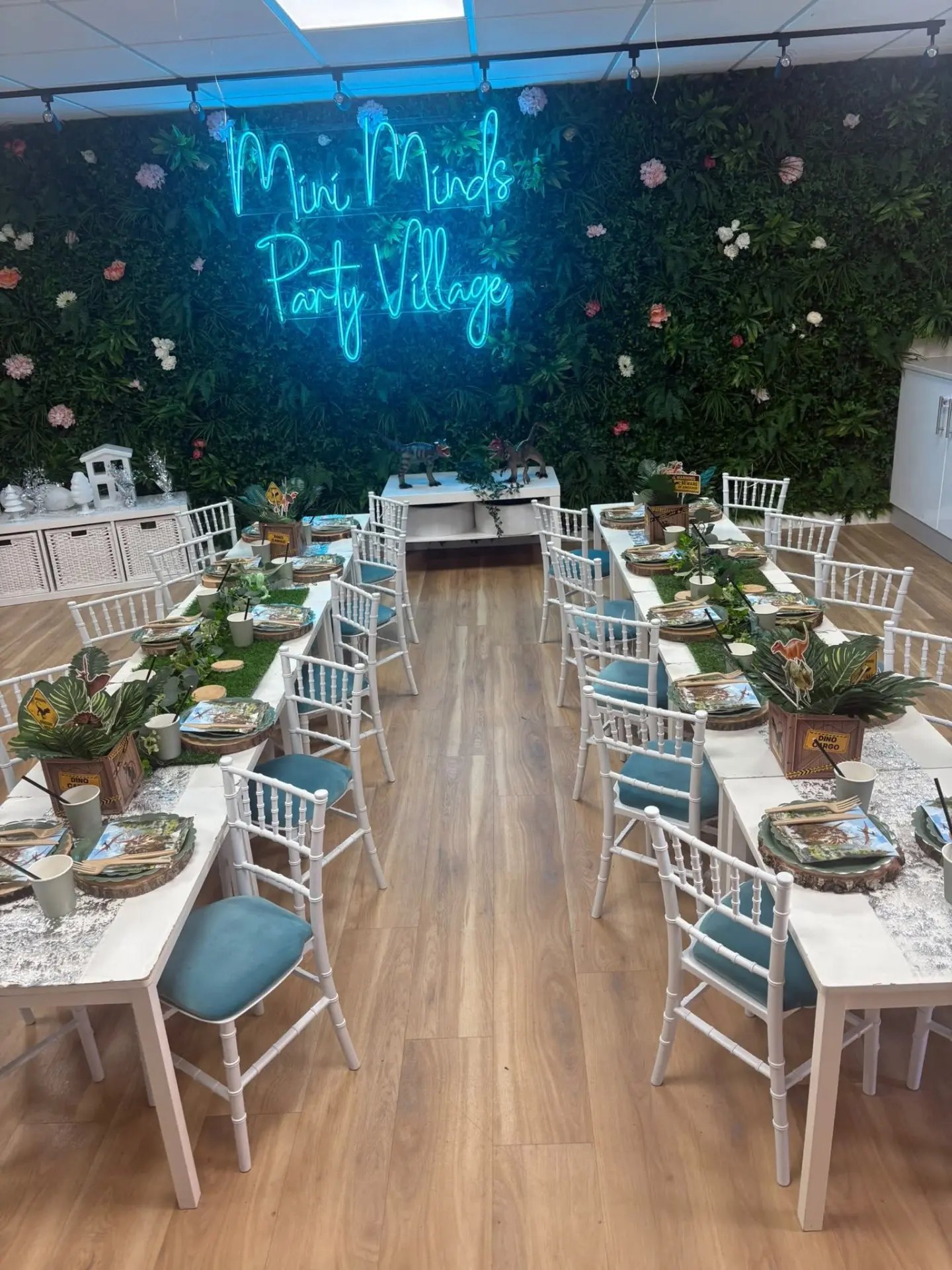 The Mini Minds party room laid out for a dinosaur party, with two long tables, white chairs and a lit Mini Minds sign on a foliage wall